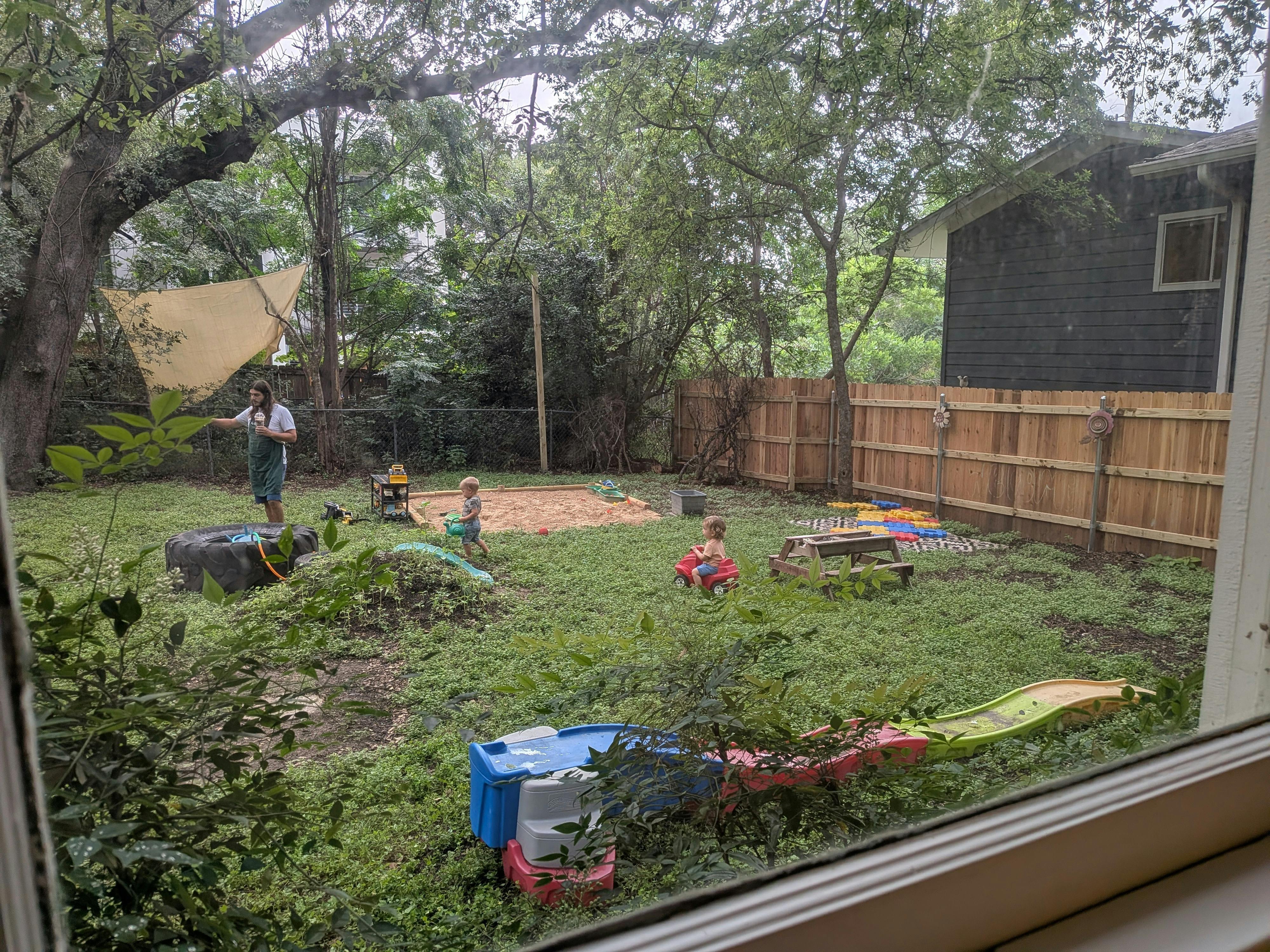 People in a green backyard with sandpit and toys