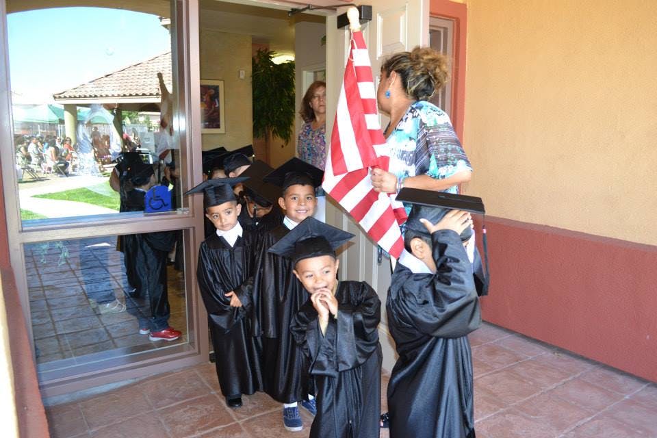 Children in graduation gowns and caps with a person holding a flag