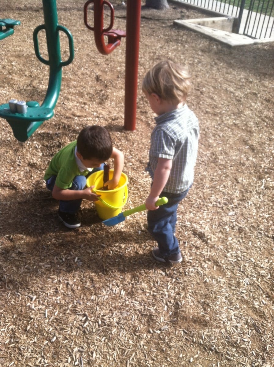 Two children playing with a yellow bucket and shovel on wood chips