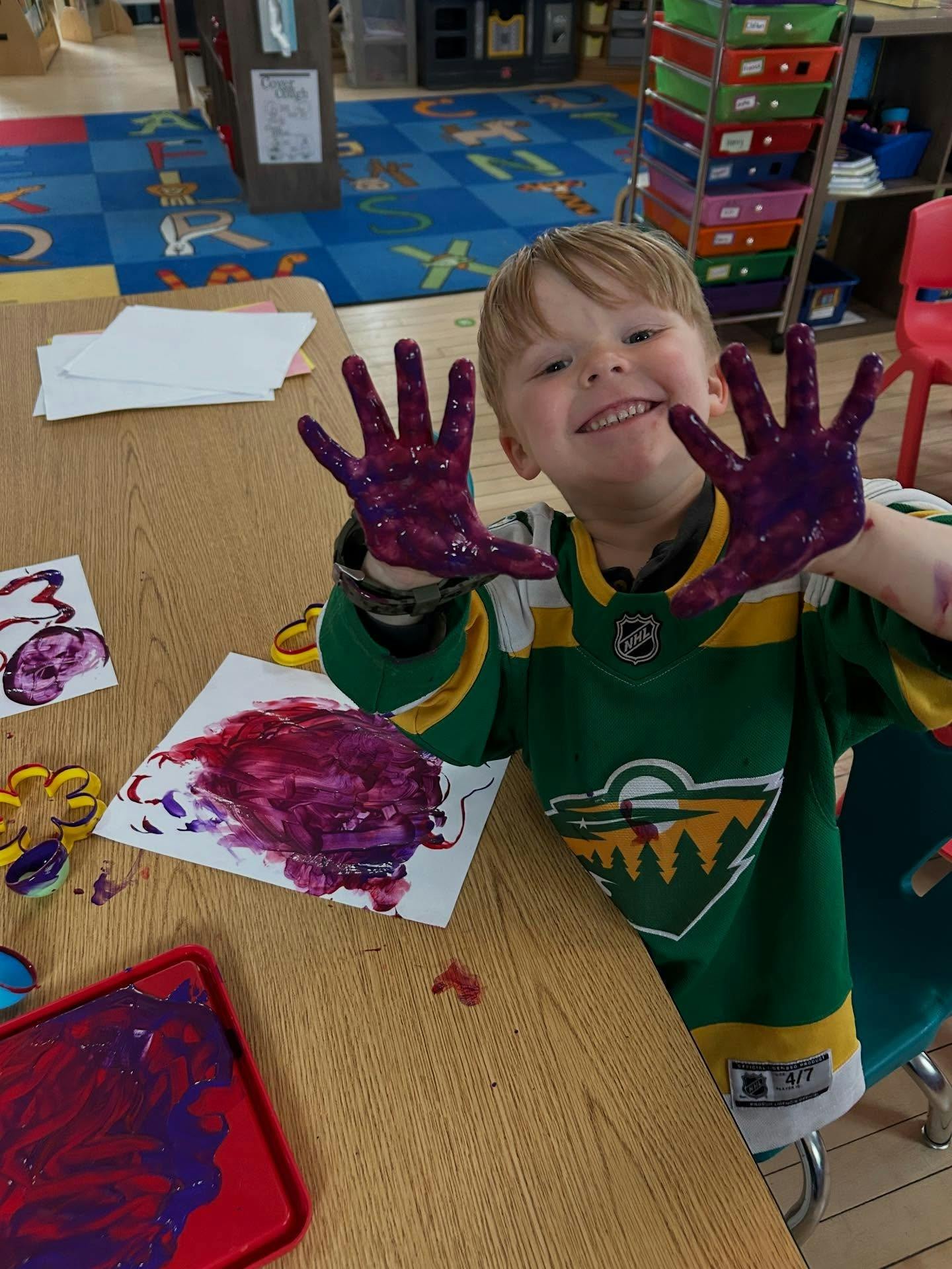 Child showing hands covered in purple paint at a table