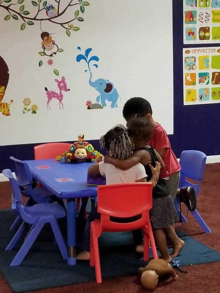 Two children embracing in a classroom with a third child seated at a table