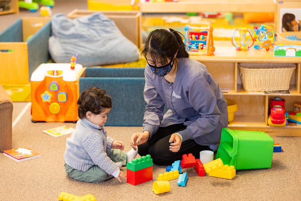 Person in mask and child playing with colorful building blocks on the floor