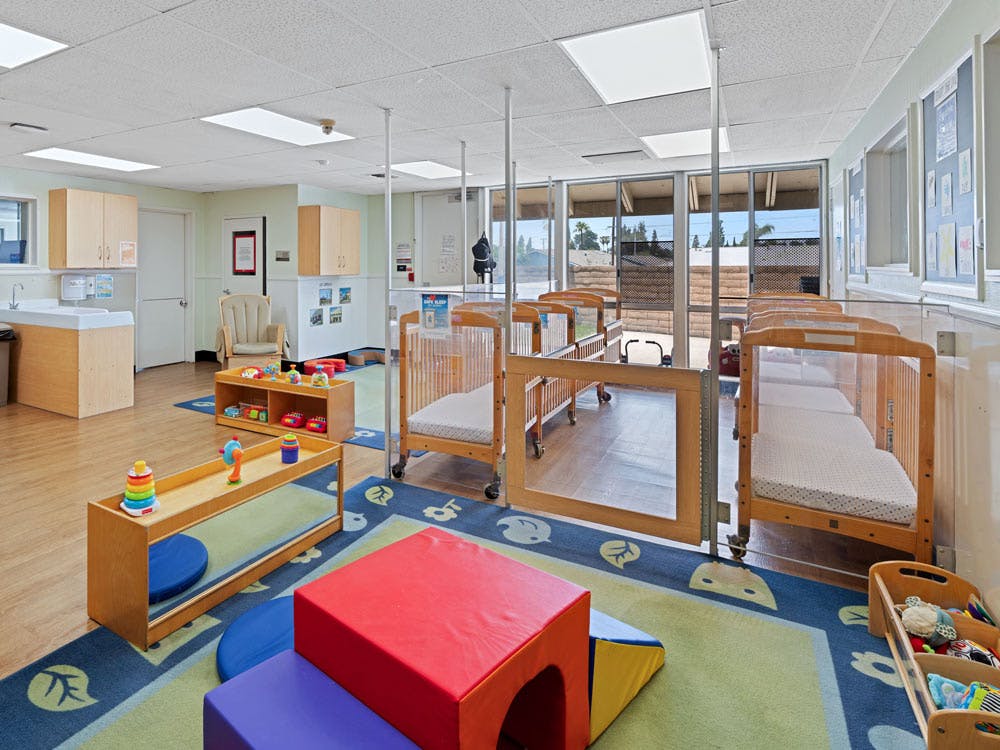 Cribs inside a daycare room with a colorful play rug and child-sized furniture