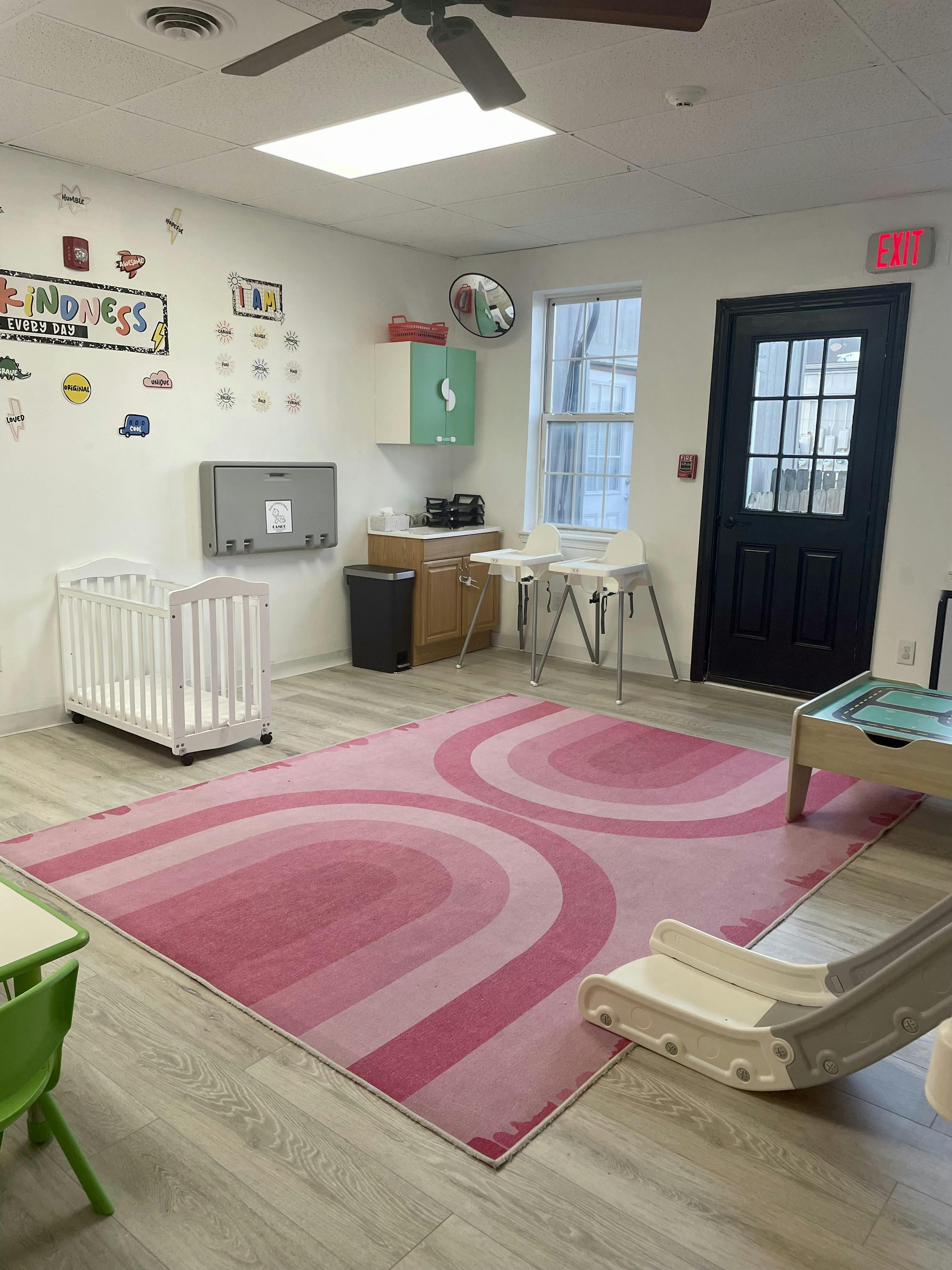 Childcare room with a crib, high chairs, and play equipment on a pink rug