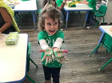 Child showing hands covered in green paint and glitter in a classroom