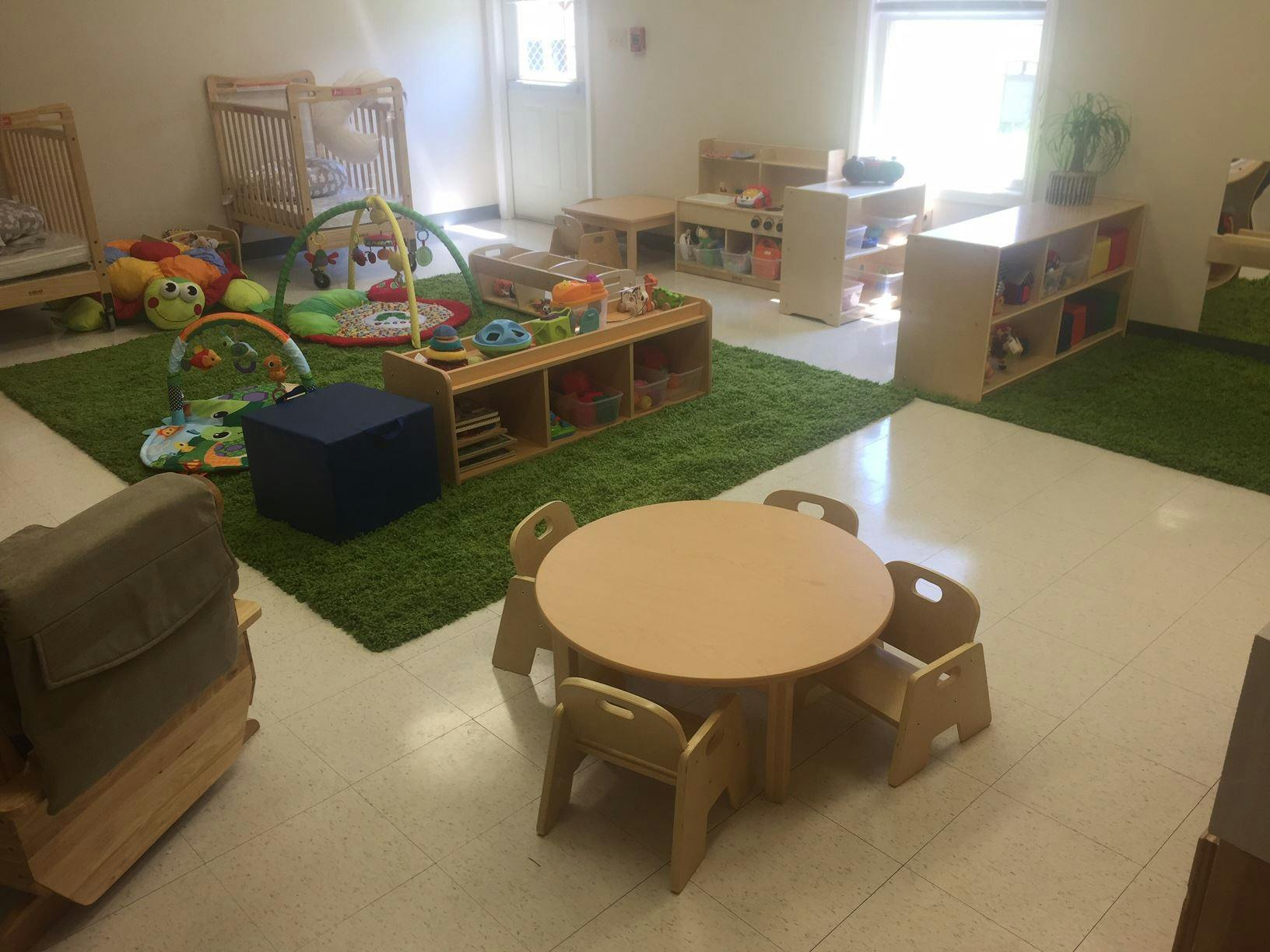 Playroom with small tables and chairs, crib, and play mats on green rugs