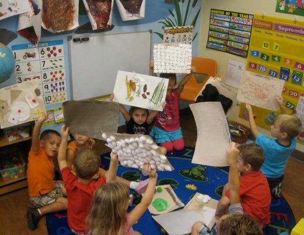 Children sitting on a rug holding up their artwork in-progress art projects
