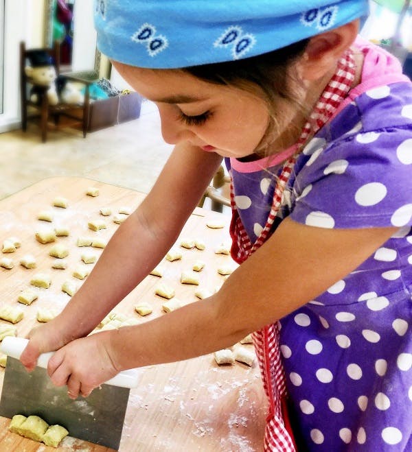 Child in an apron using a dough cutter on a wooden surface with small pieces of dough