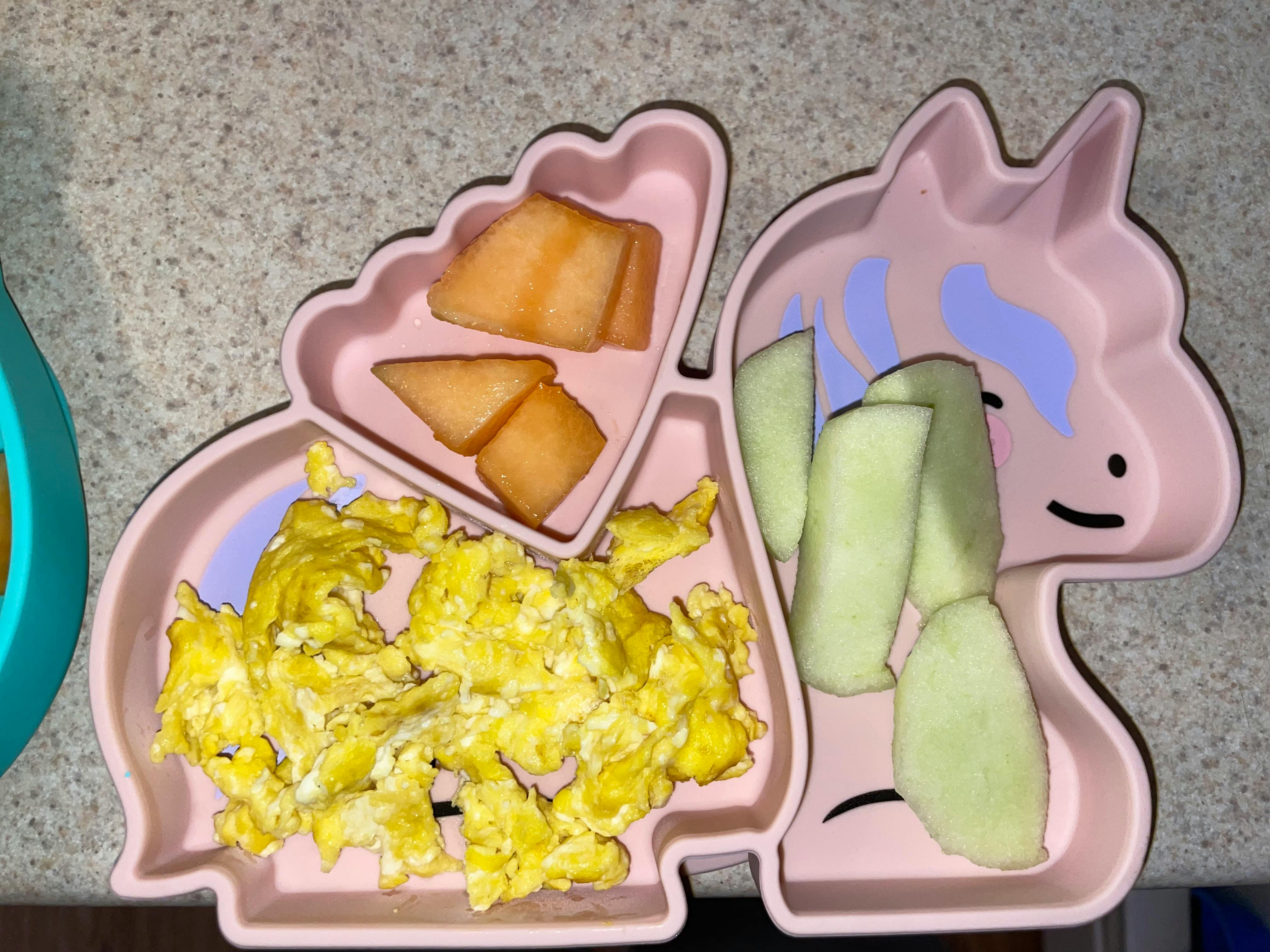 A divided pink plate in the shape of a unicorn with scrambled eggs and fruit