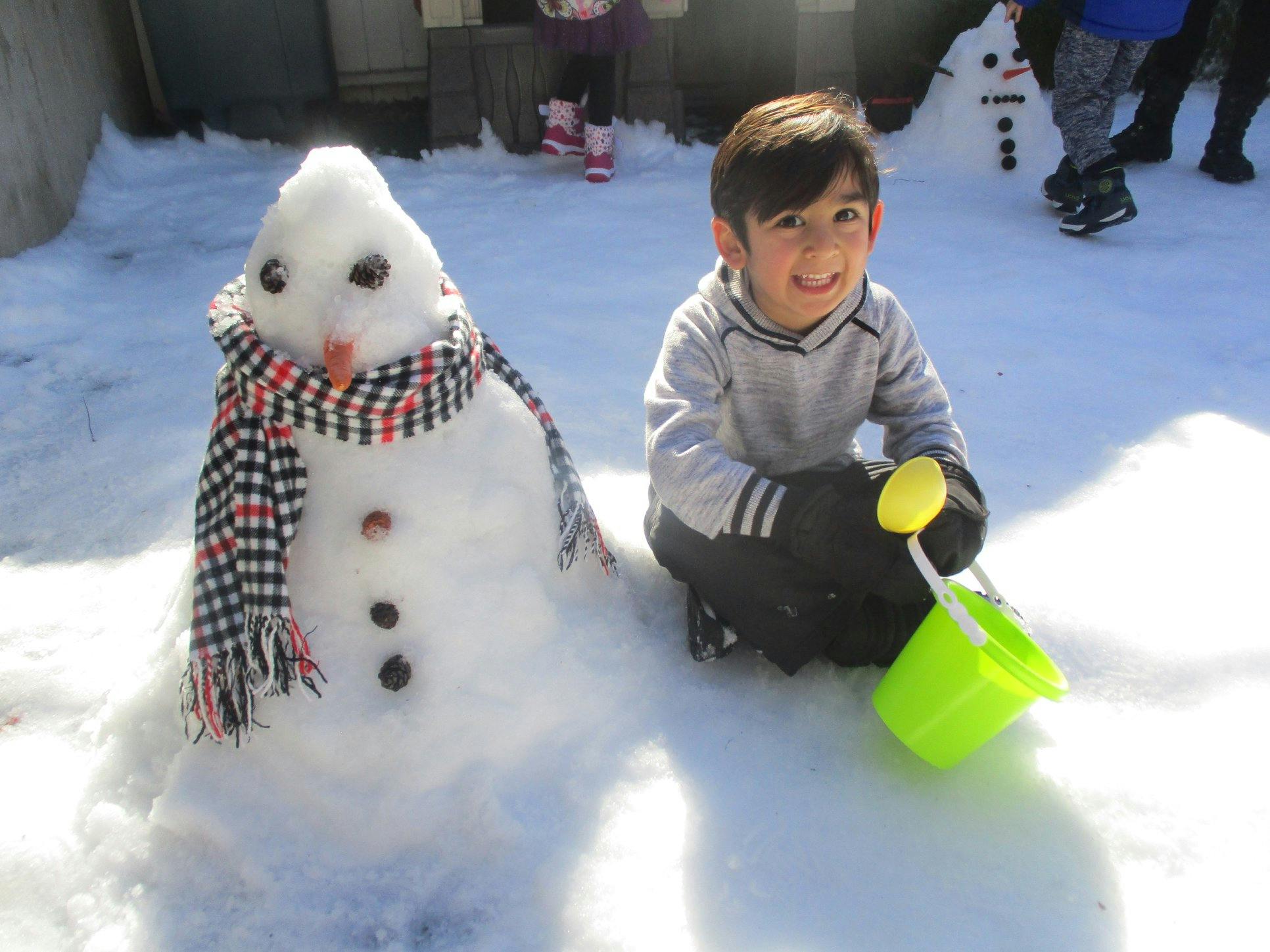 Child kneeling in snow next to a snowman, holding a toy shovel and bucket