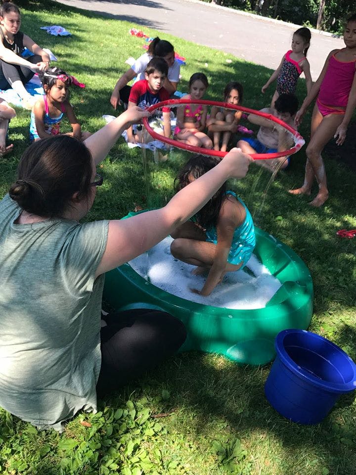 Person holding a large bubble wand over a child in a bubble-filled tub