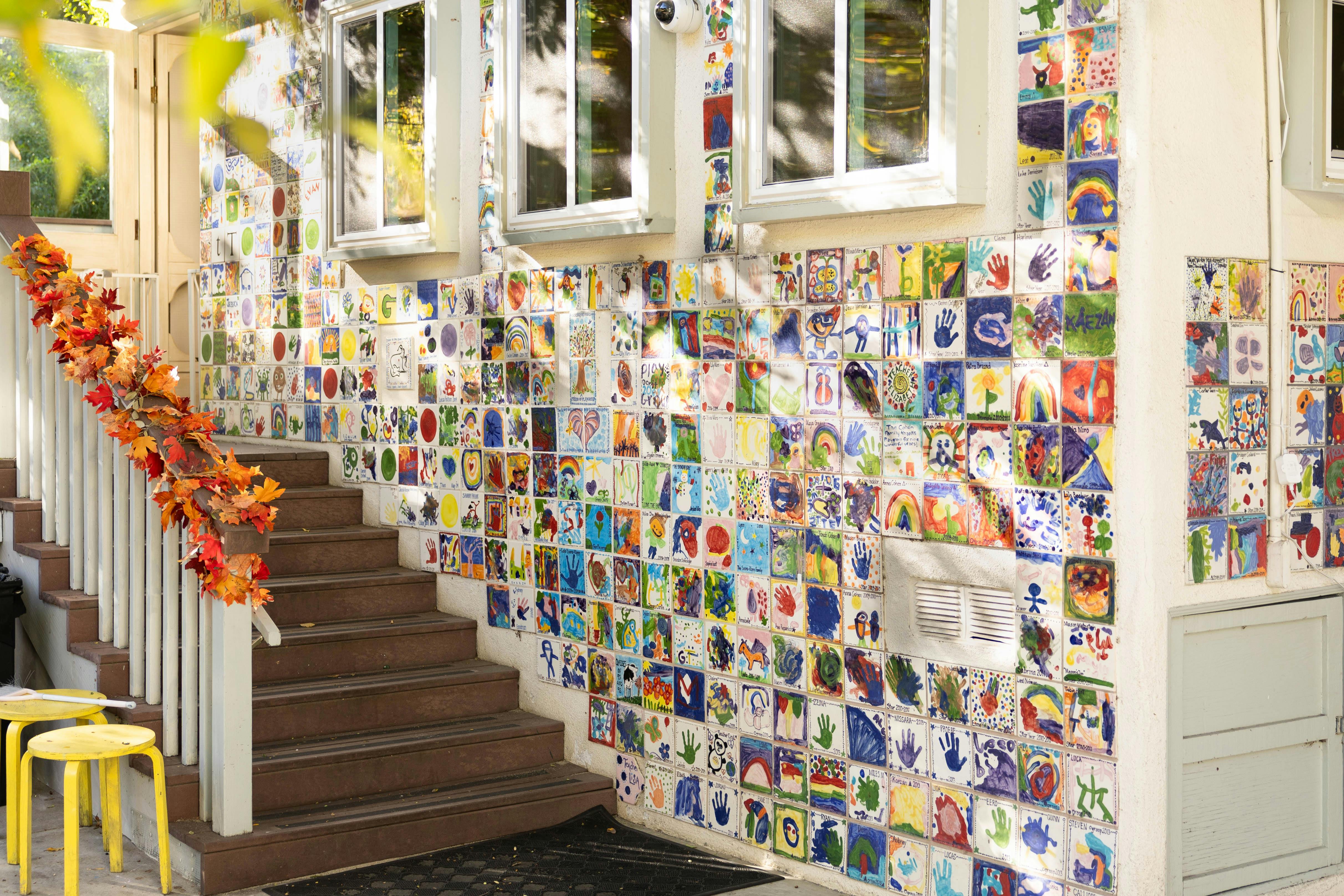 A wall covered in colorful child-created tiles next to outdoor stairs with fall leaf garland