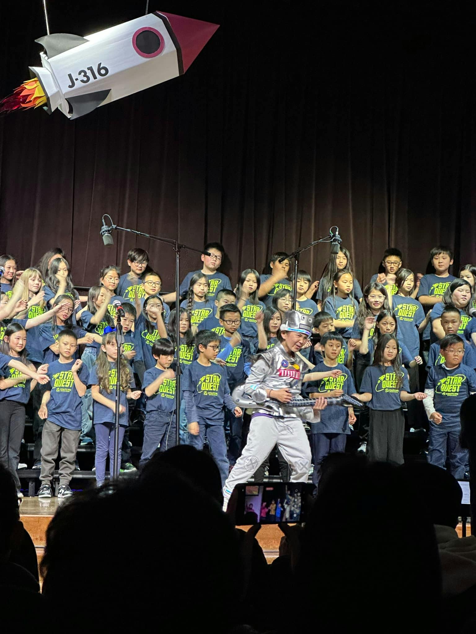 Children on stage with a person in a silver costume holding a guitar