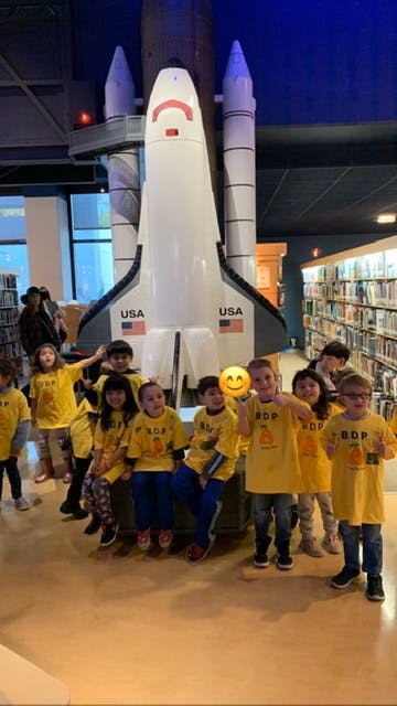 Children in yellow shirts posing in front of a large model space shuttle
