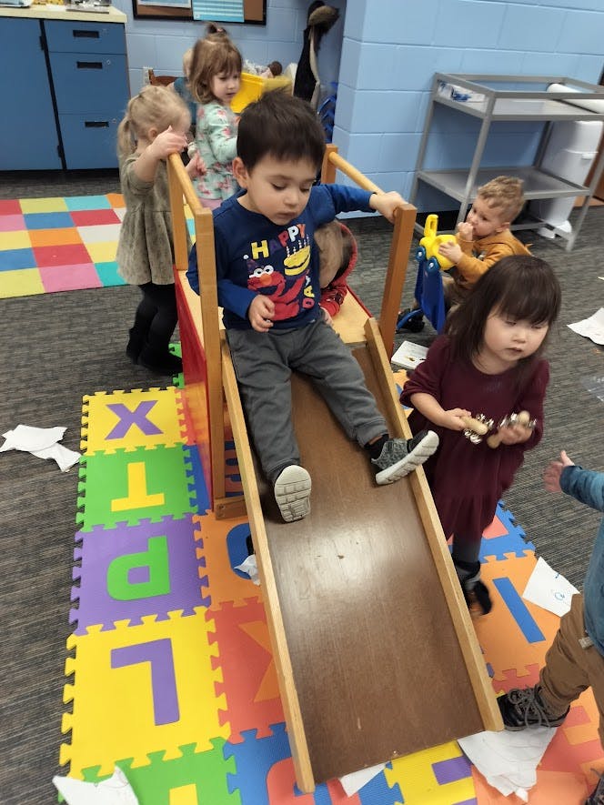 Child sliding down a small wooden slide in a classroom with other children