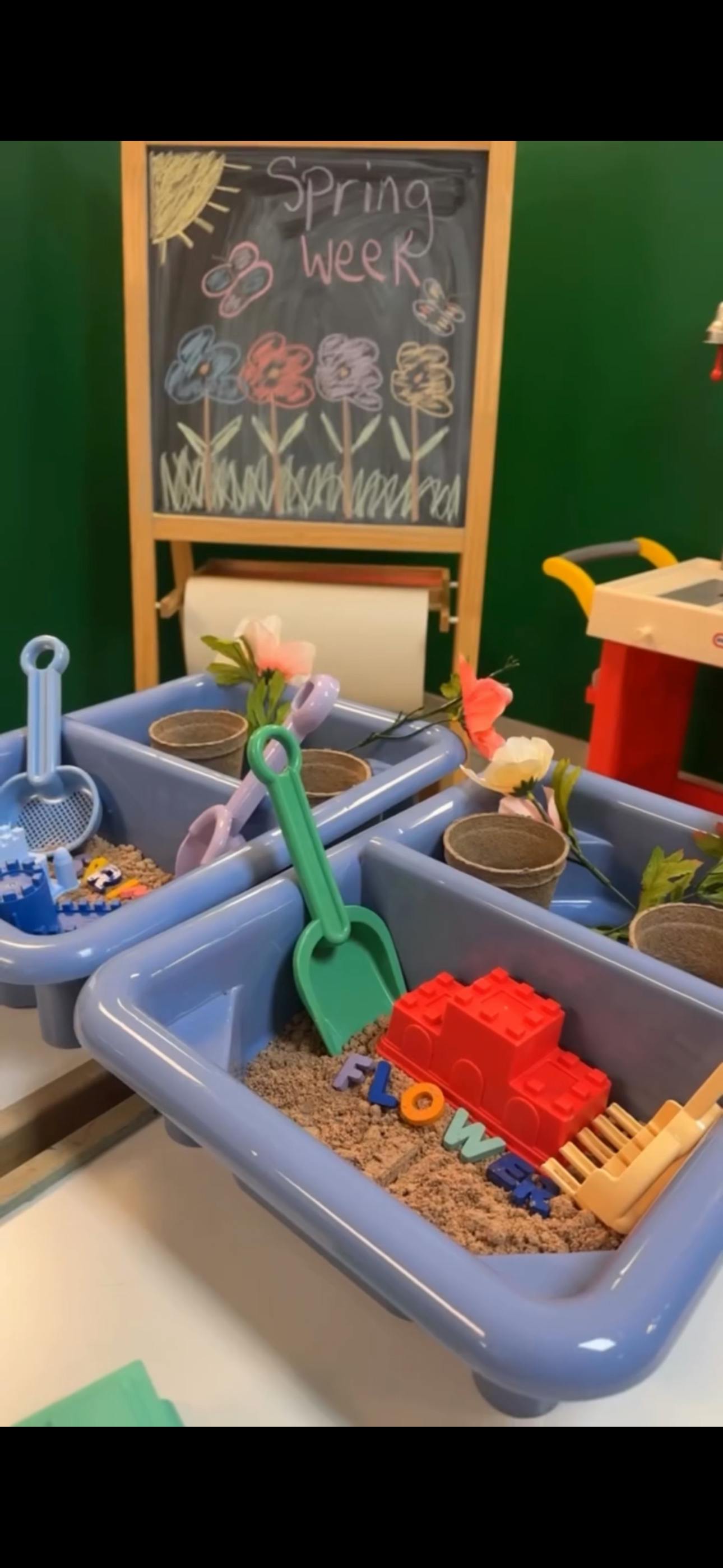 Children's sensory bins with sand, plastic toys, and a "Spring week" chalk sign