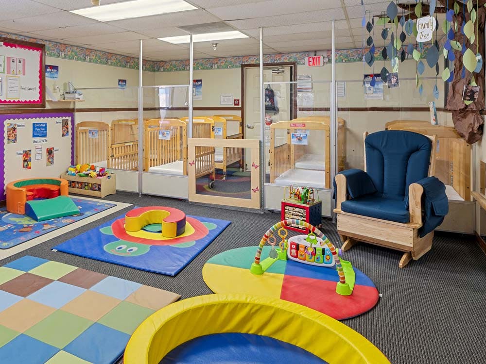Cribs separated by clear partitions in a daycare room with playmats and a rocking chair