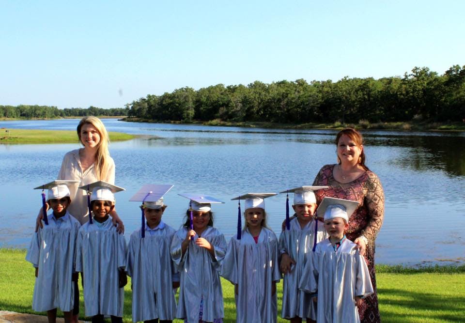 Two people with children in graduation caps and gowns by a lake