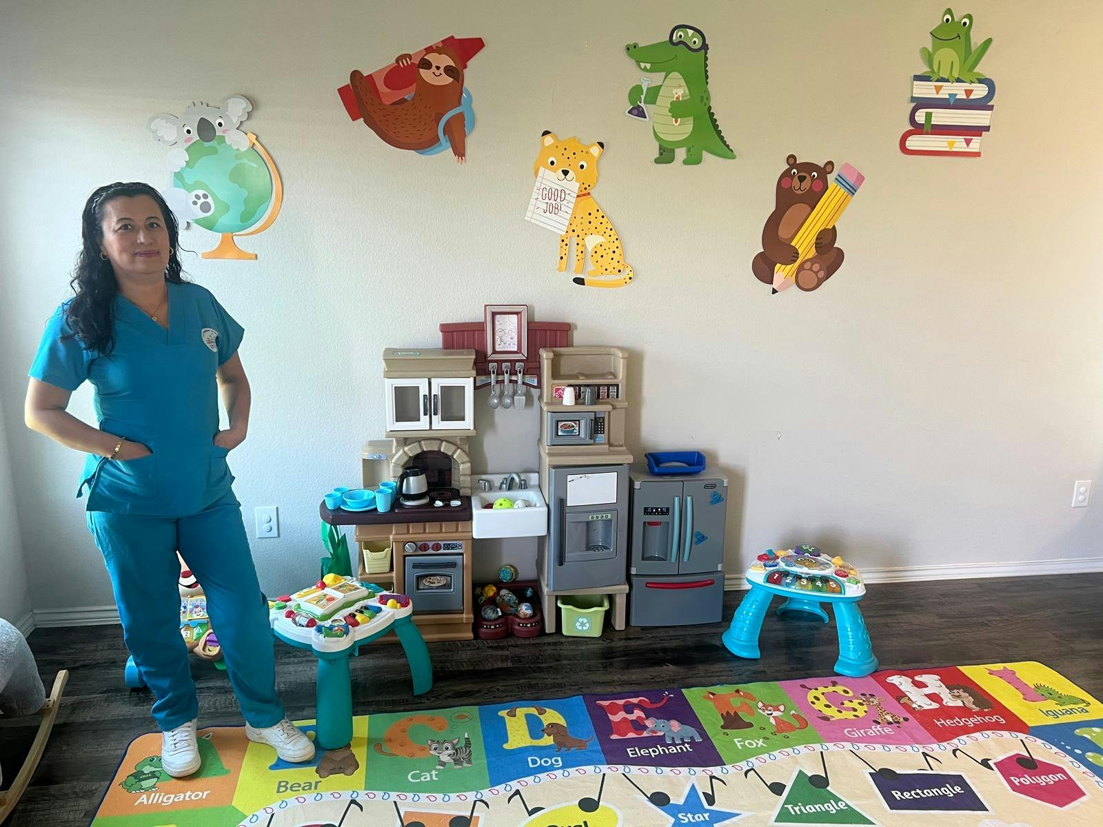 Person in teal scrubs standing in a play area with toy kitchen and alphabet rug