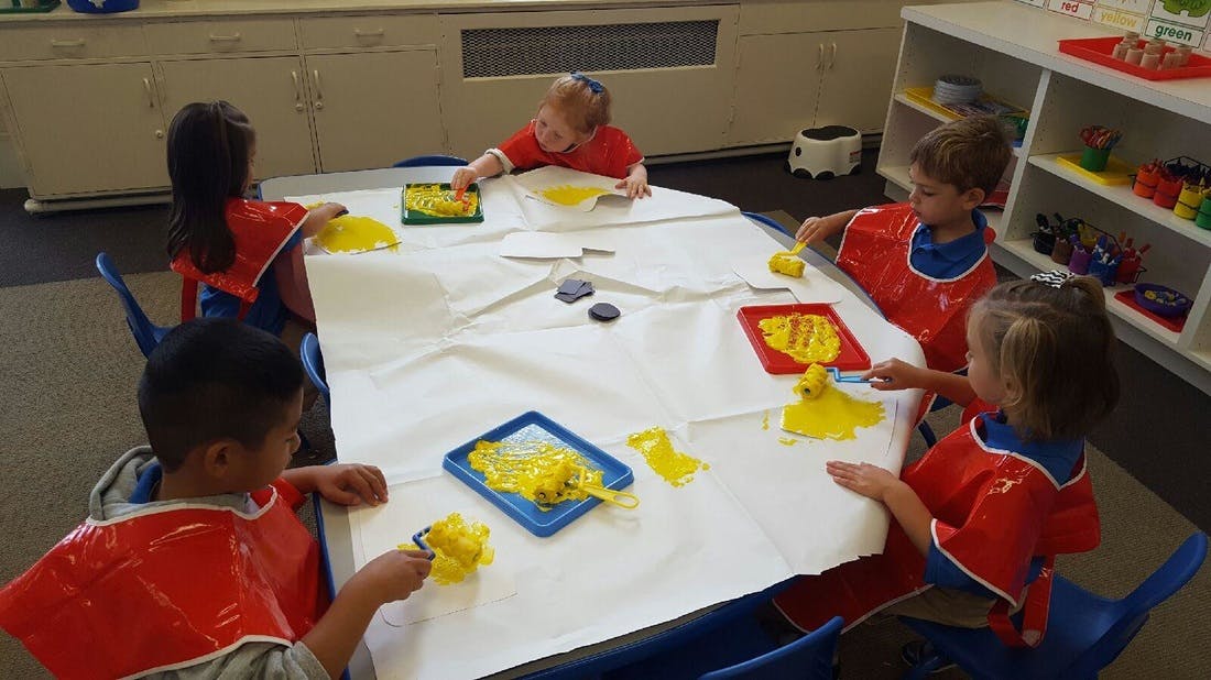 Children in red smocks painting with yellow paint at a table