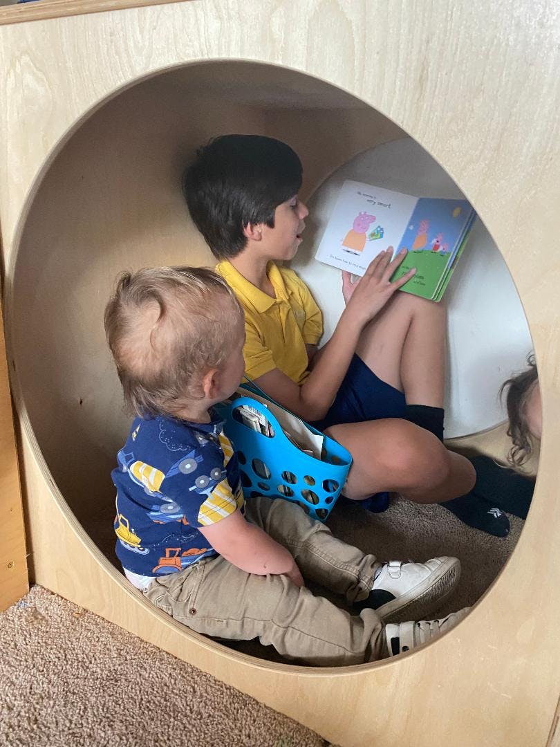 Two children sitting in a wooden cubby, one reading a book