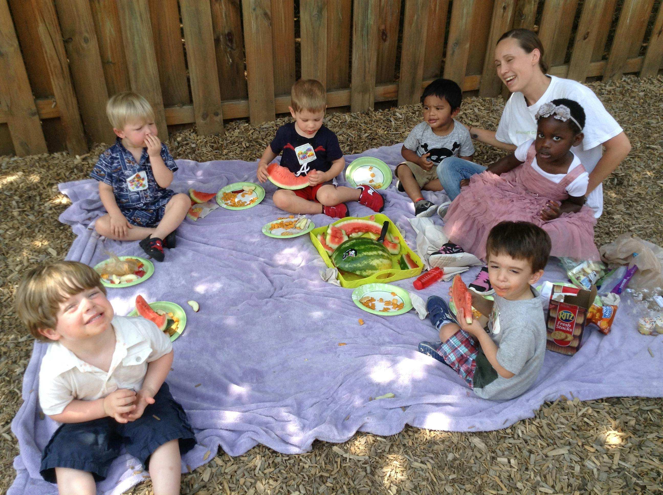 Group of children and an adult sitting on a blanket eating watermelon