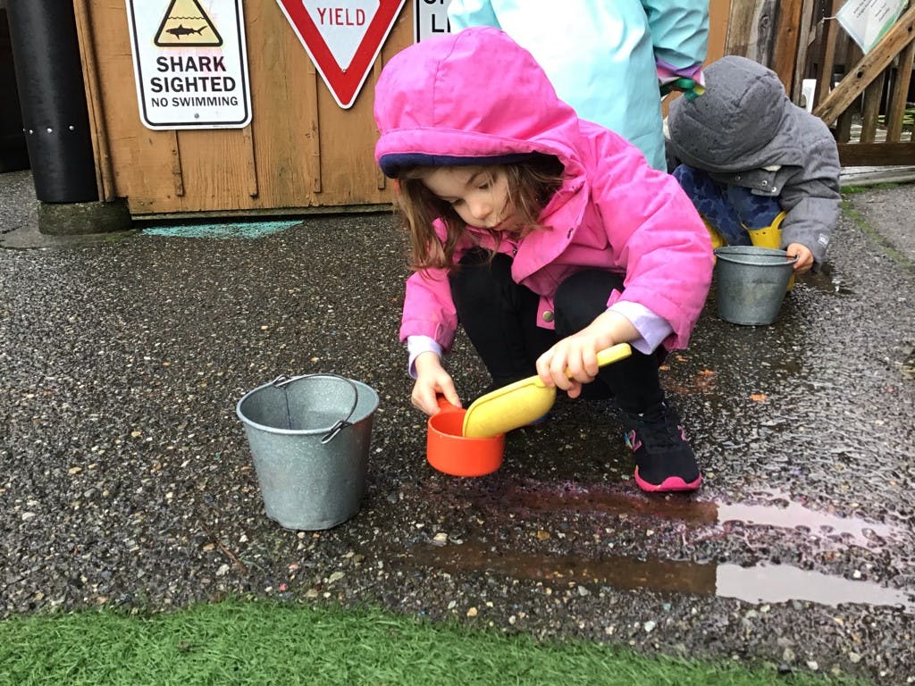 Child in pink jacket scooping water from a puddle into an orange cup
