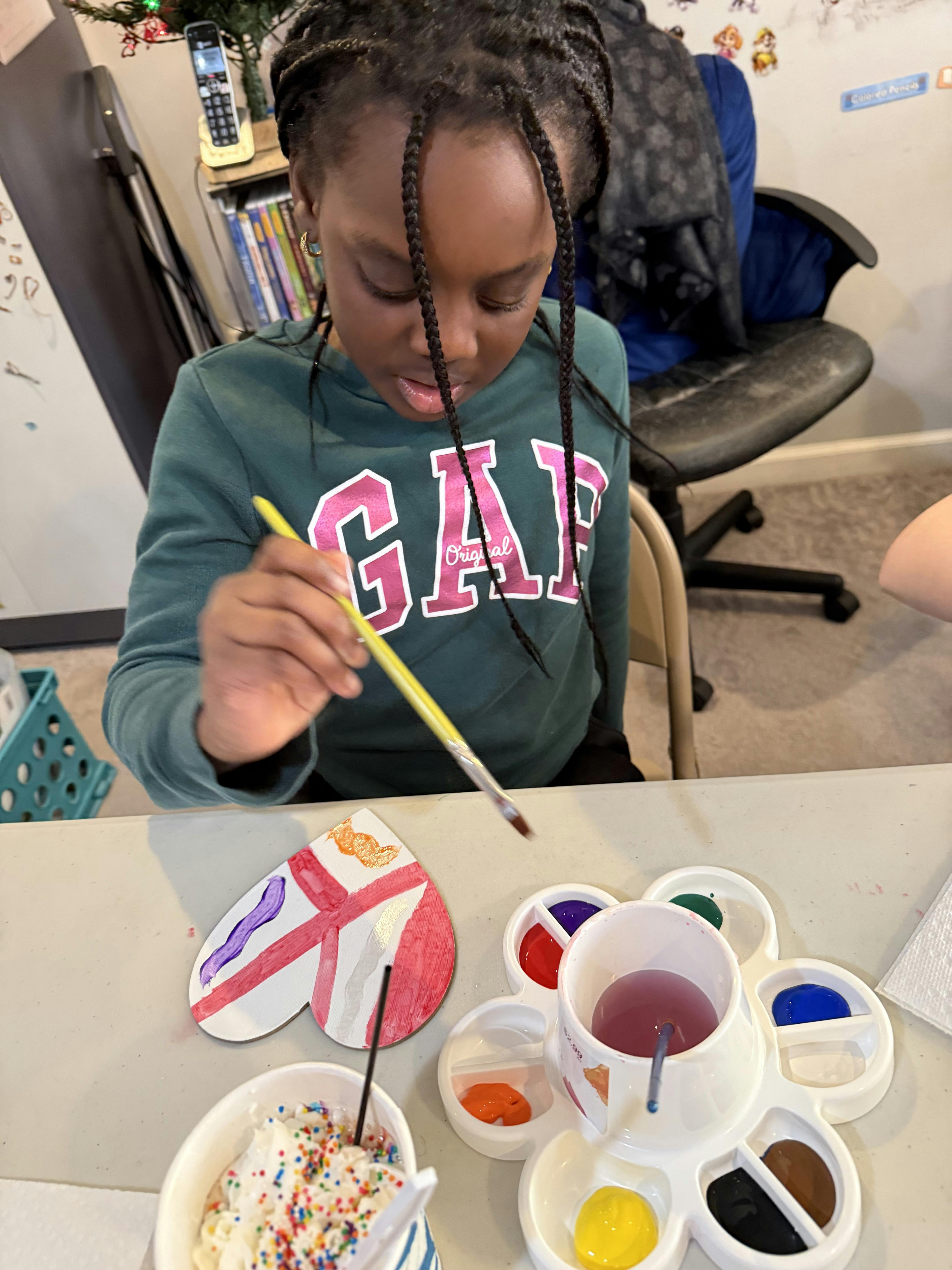 Child painting a heart-shaped object on a table next to a paint palette