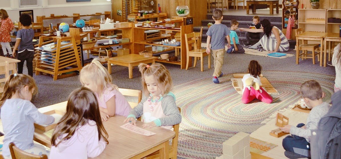 Children in a classroom, some seated at tables, others on the floor