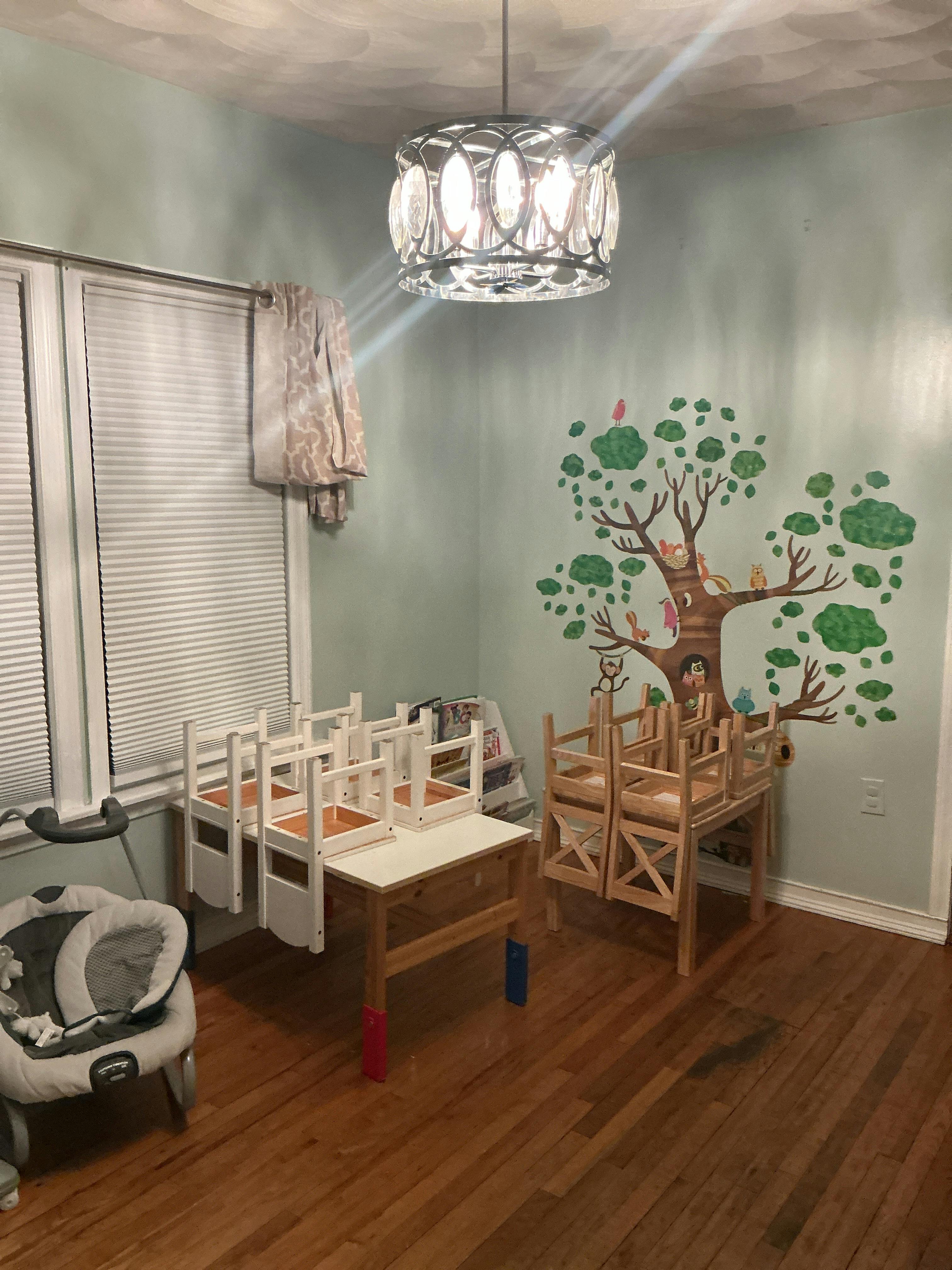 Room with tree decal on wall, wooden and white tables, chairs, and baby bouncer