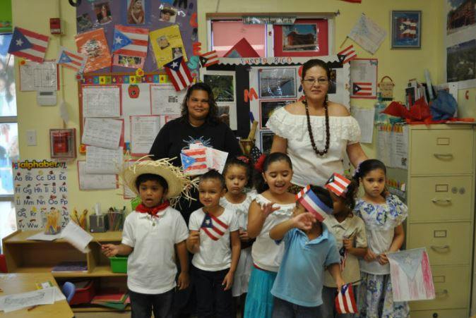 Two people and multiple children holding small flags in a classroom