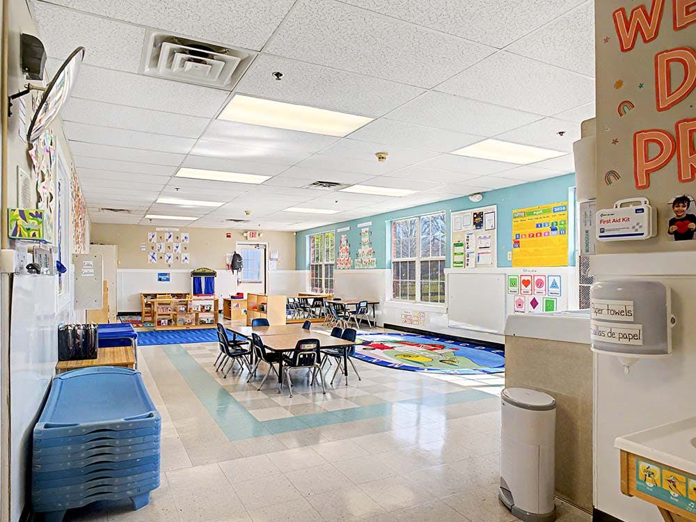 An empty classroom with small tables and chairs, and sleeping mats stacked mats