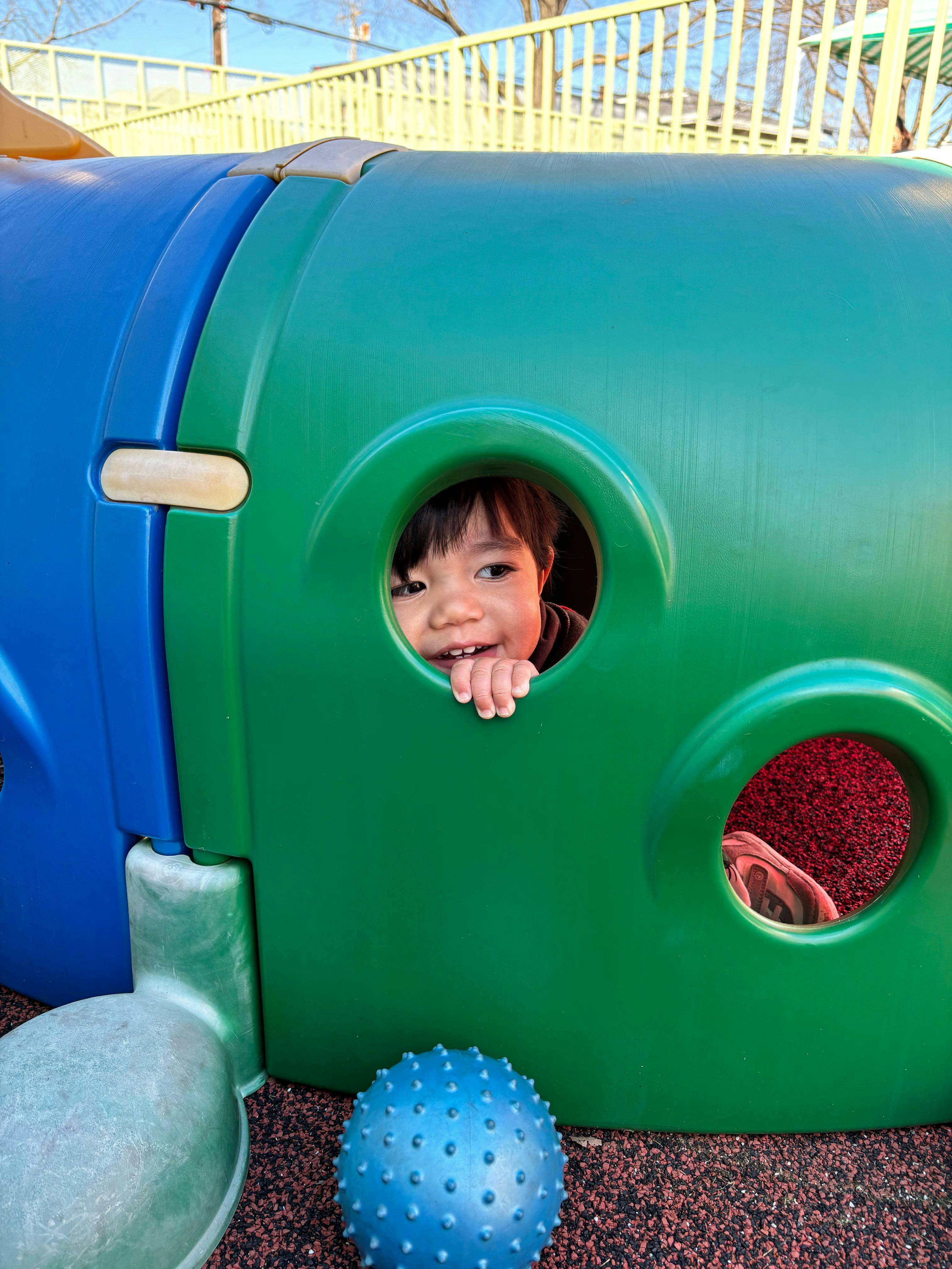 Child looking out of a hole in a green playground tunnel