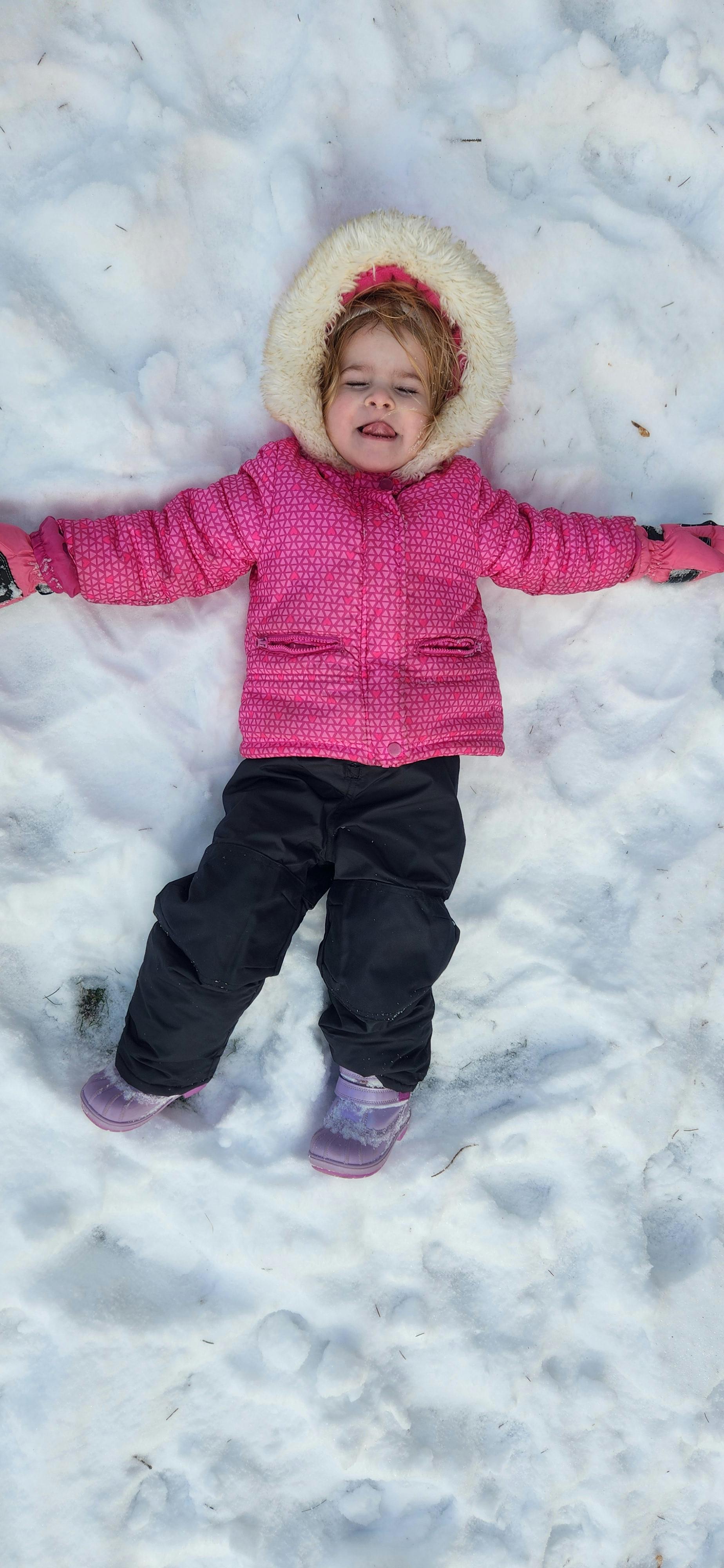 Child in a pink coat and black pants lying in the snow