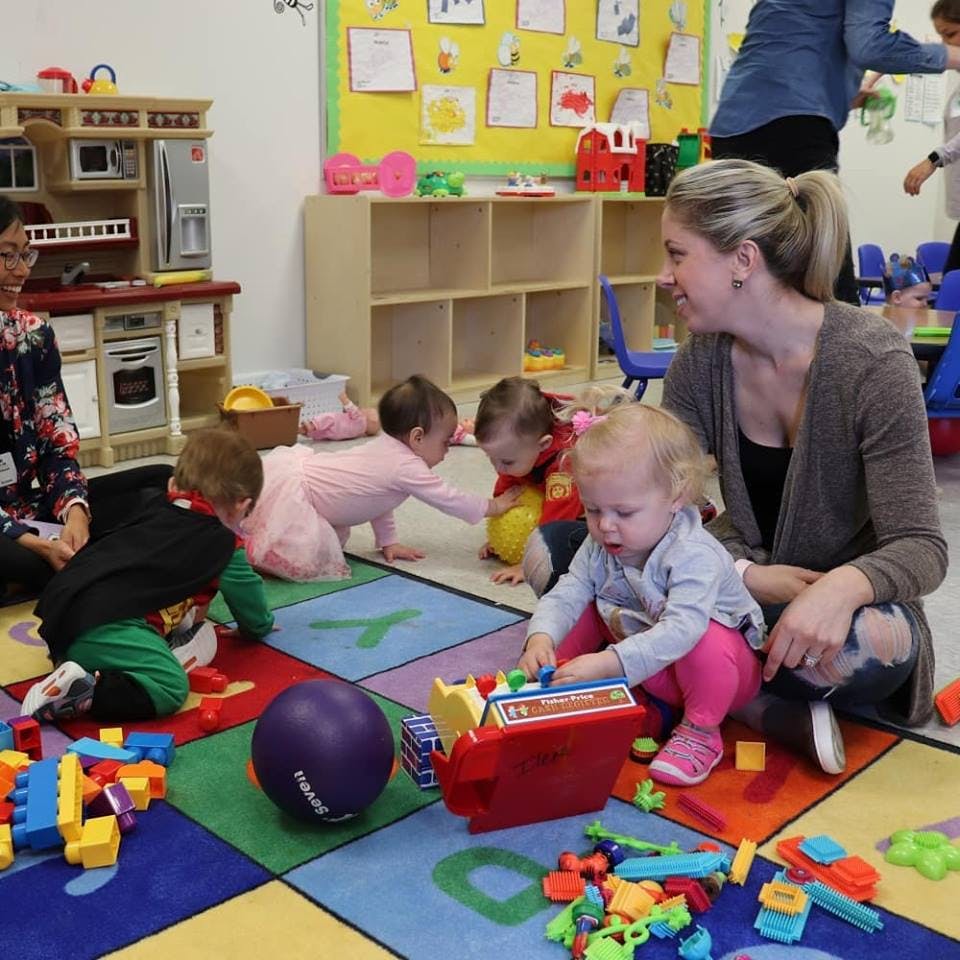 Adults and children playing with toys on a colorful rug in a classroom