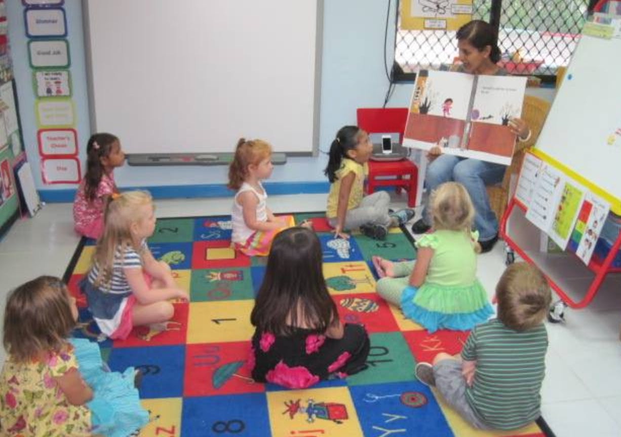 Adult reading a book to children sitting on a colorful rug in a classroom