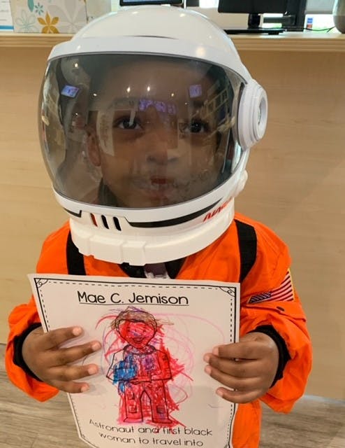 Child in astronaut helmet and orange suit holding a drawing of Mae C. Jemison