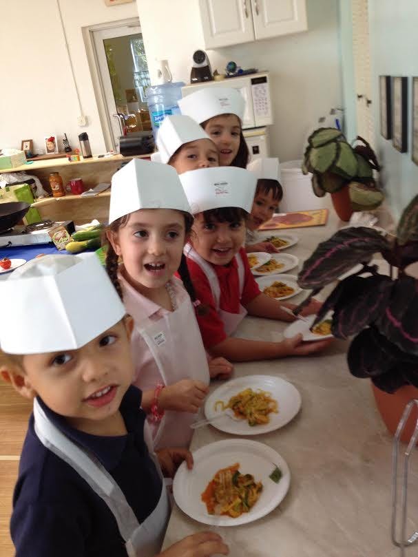 Children in chef hats and aprons holding plates of food in a kitchen
