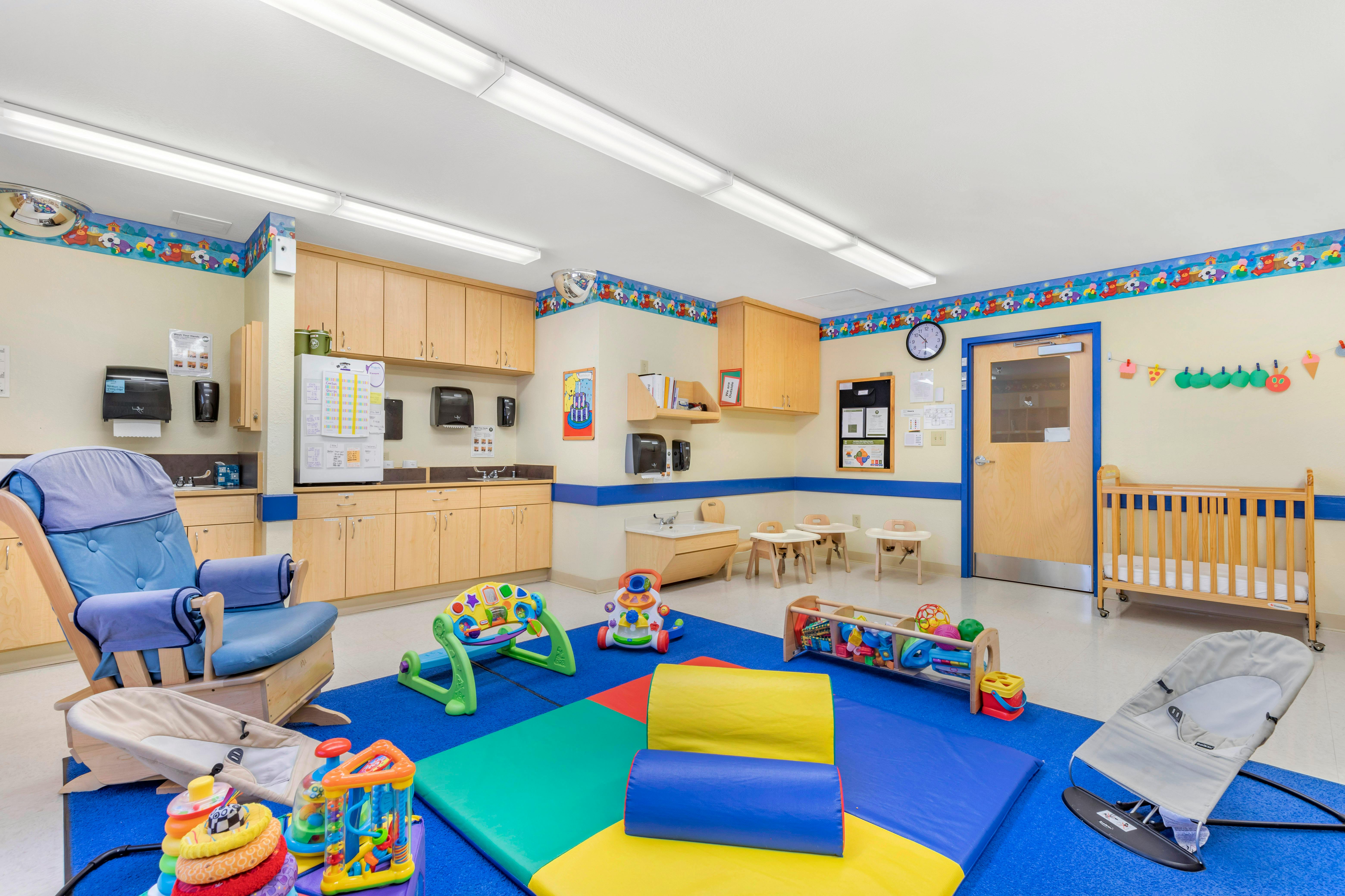 A childcare room with a crib, rocking chair, colorful play mats, and toys