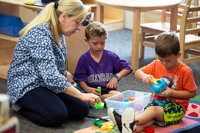 Person and two children playing with toys on the floor in a classroom