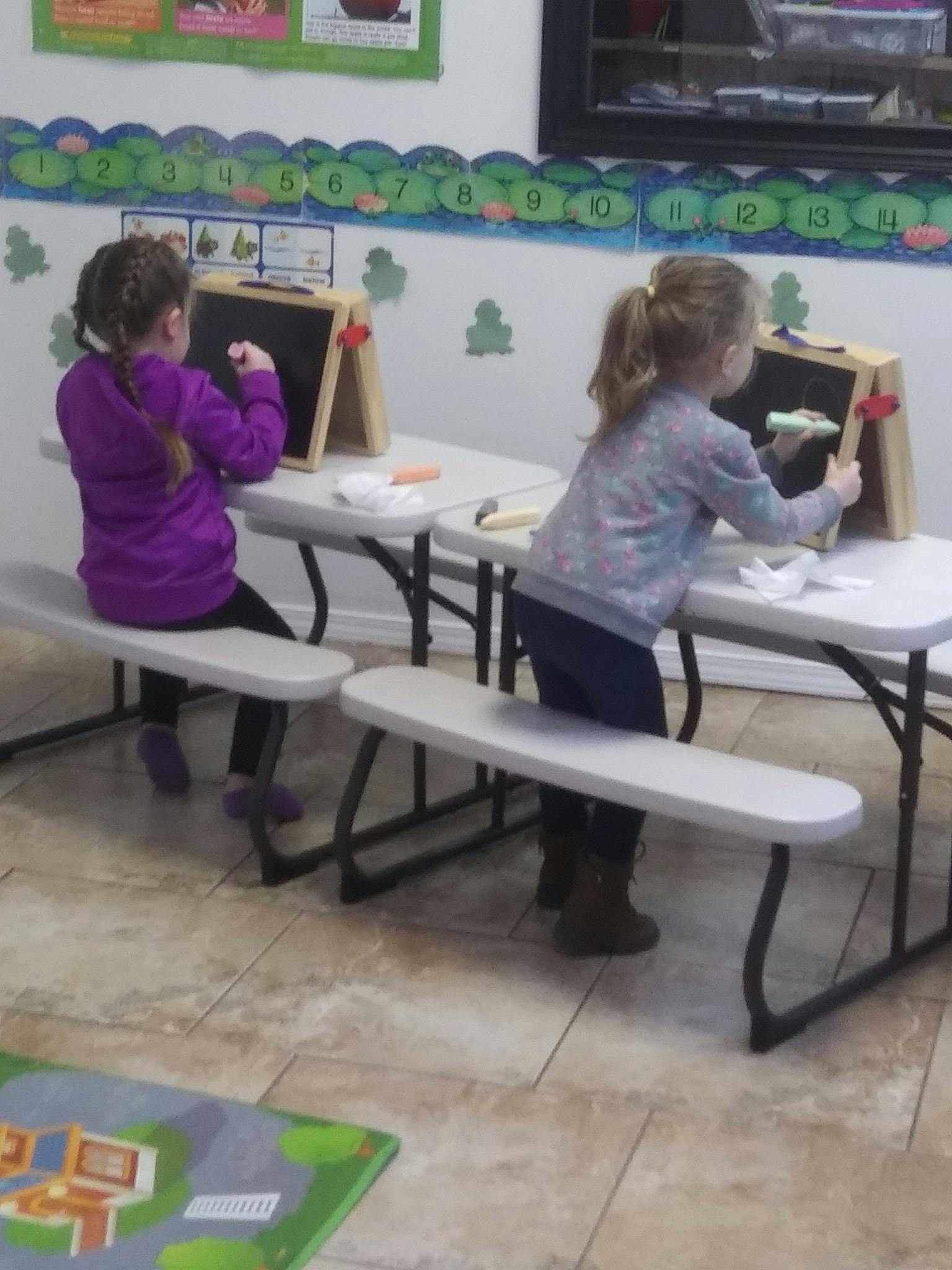 Two children drawing on chalkboards at tables