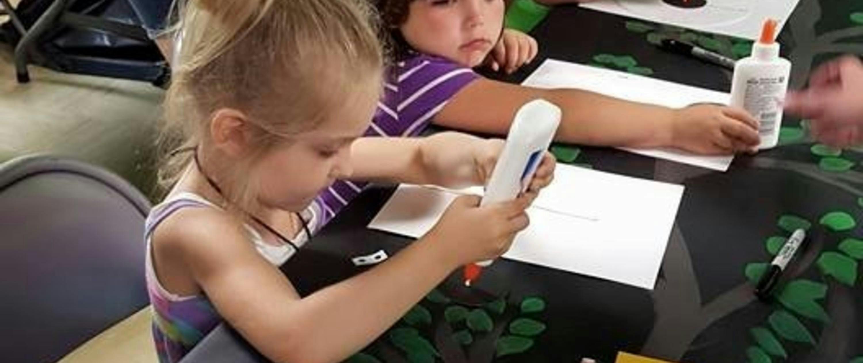 Child holding a glue bottle over paper, another child next to them