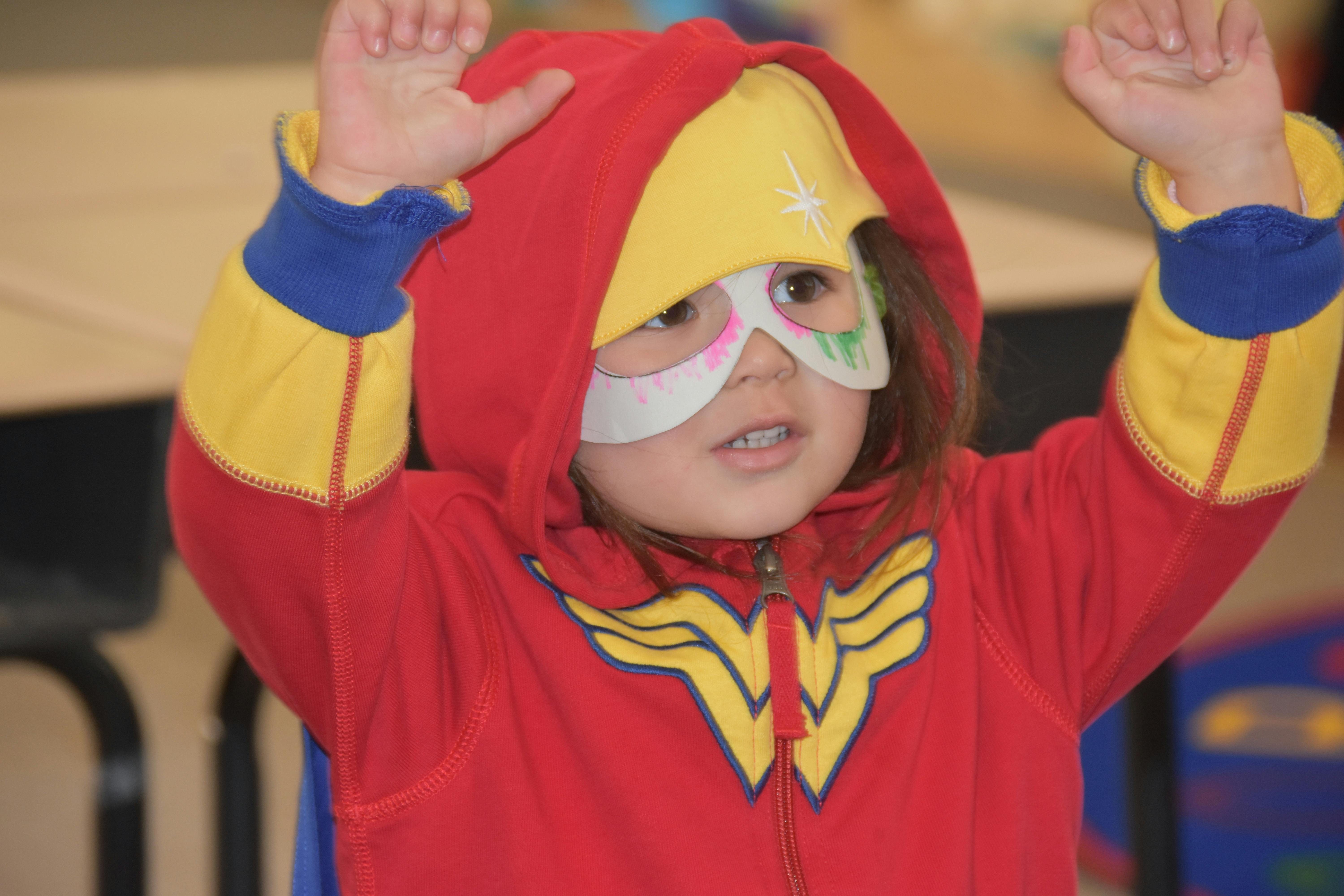 Child in a red hooded costume with a yellow emblem and a white mask, arms raised