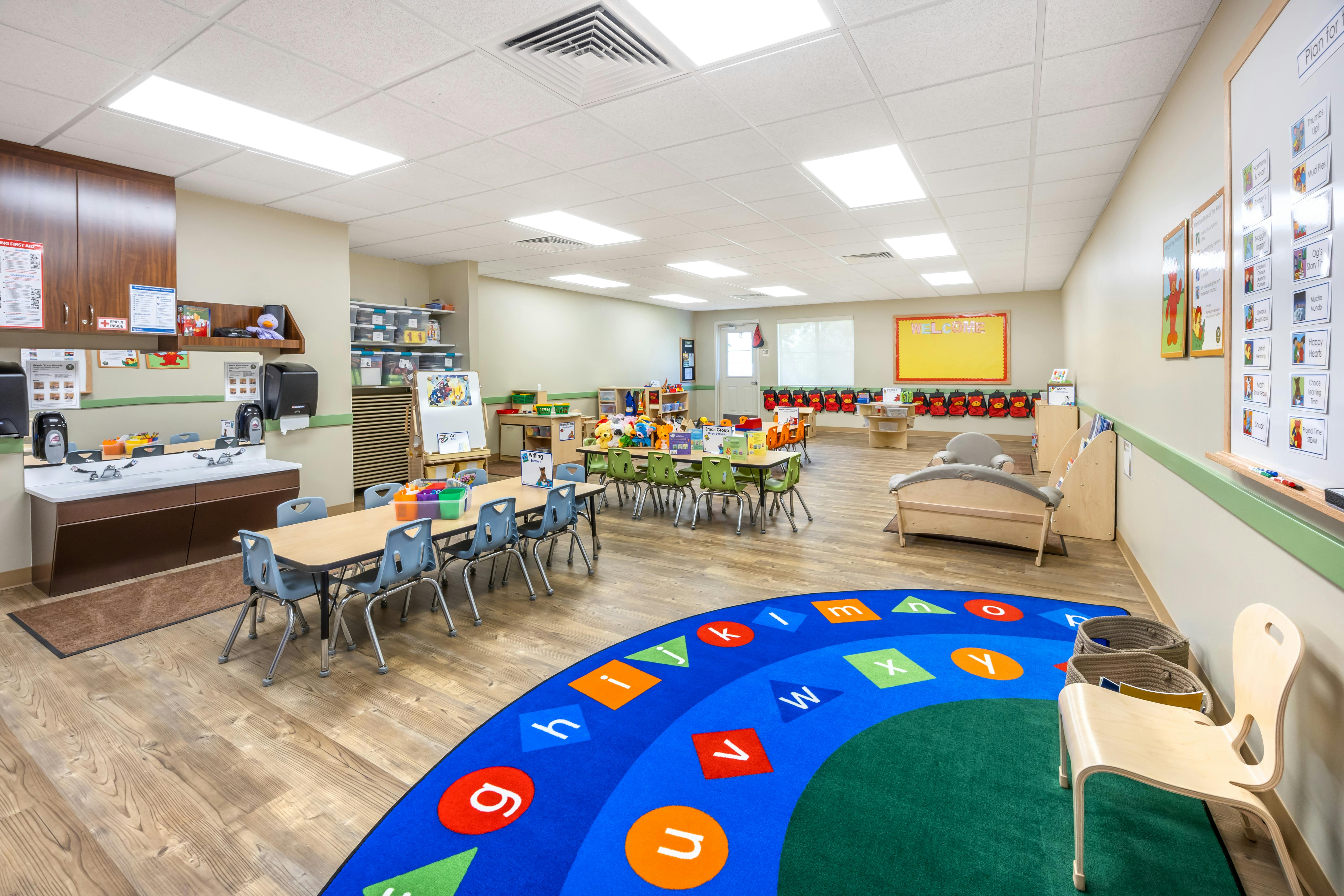 Classroom with tables, chairs, a sink area, and a colorful alphabet rug
