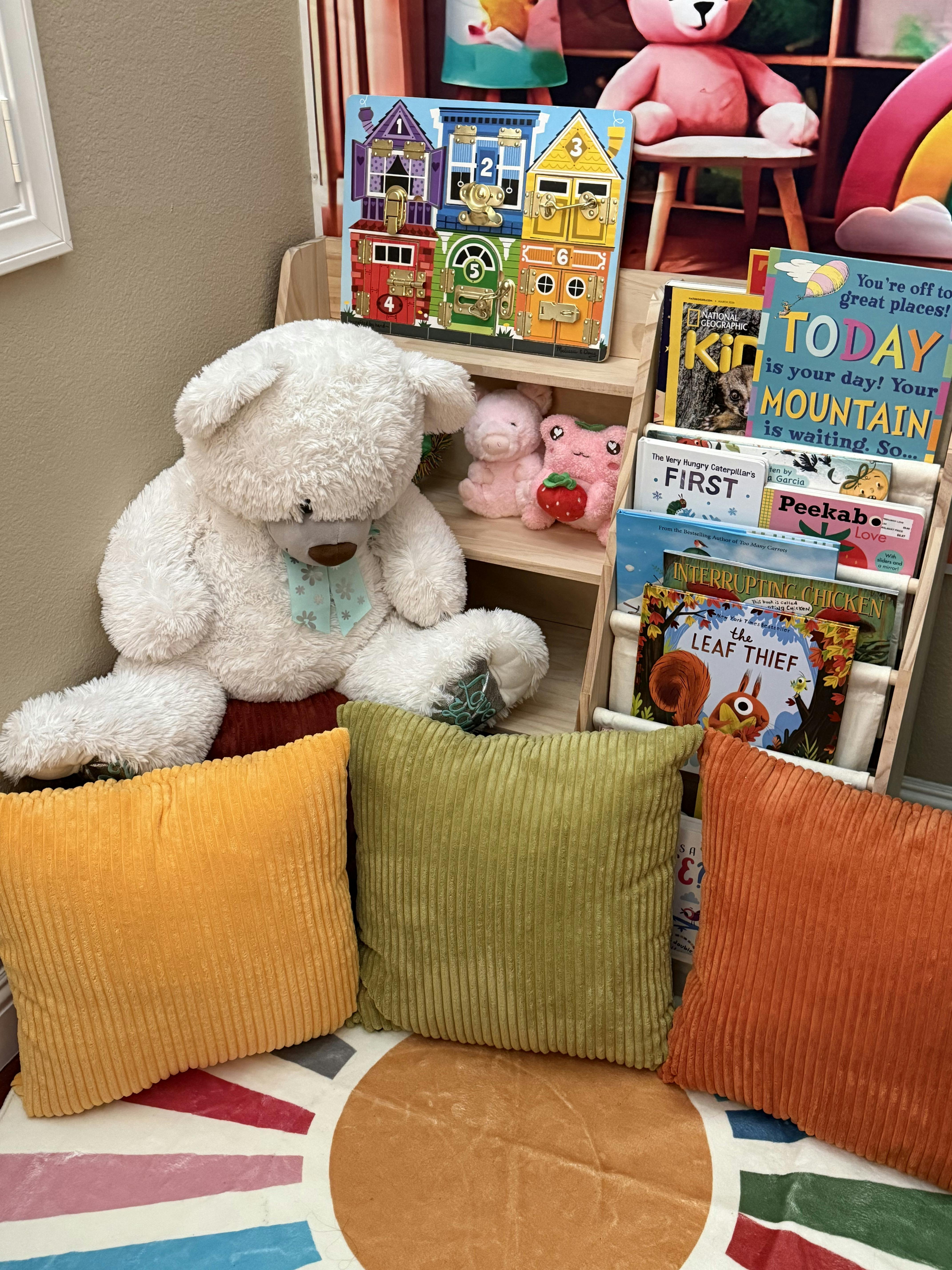 A large white teddy bear sitting next to a bookshelf filled with children's books and toys