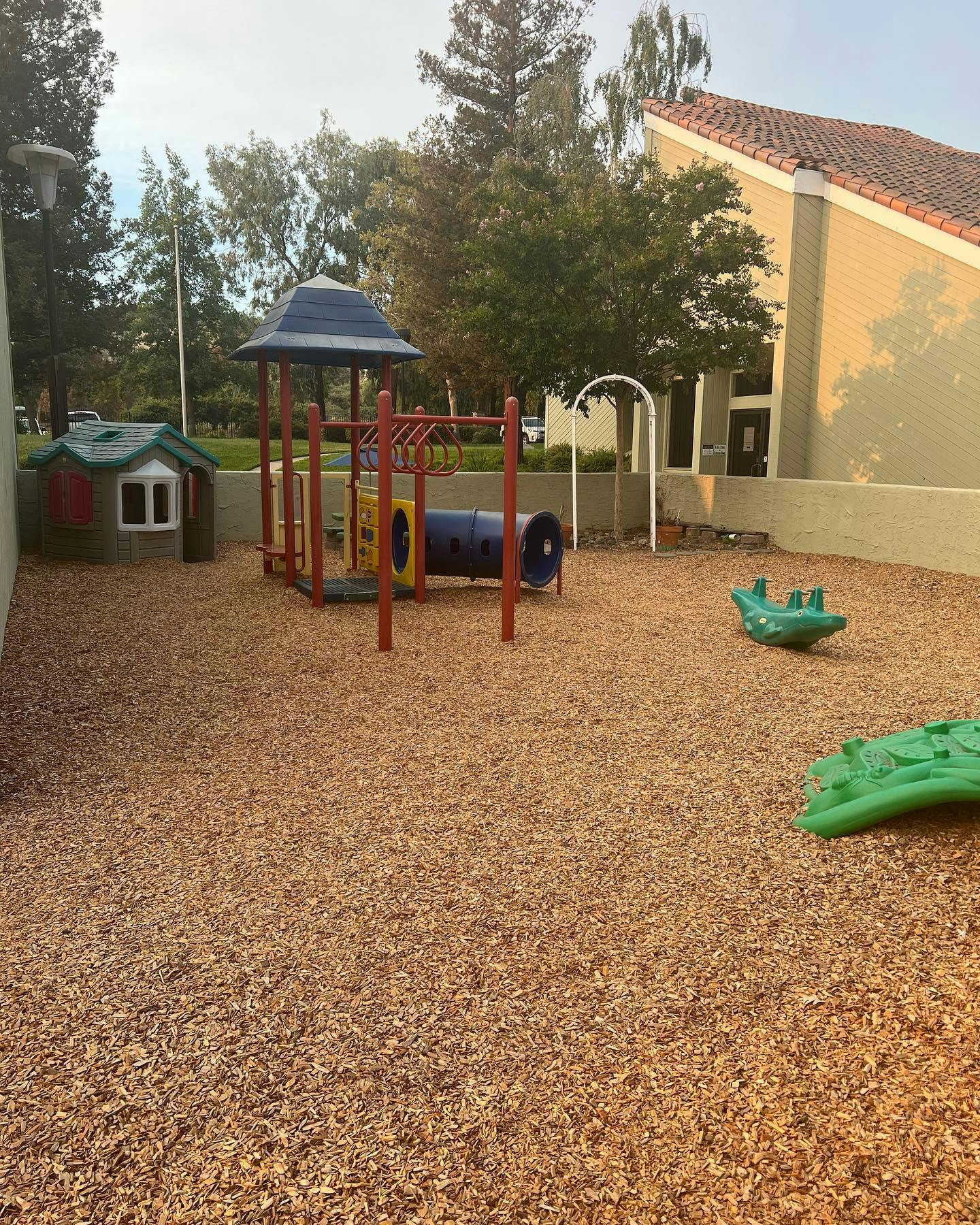Playground with wood chips, small house, climbing structure, and green rocking toys