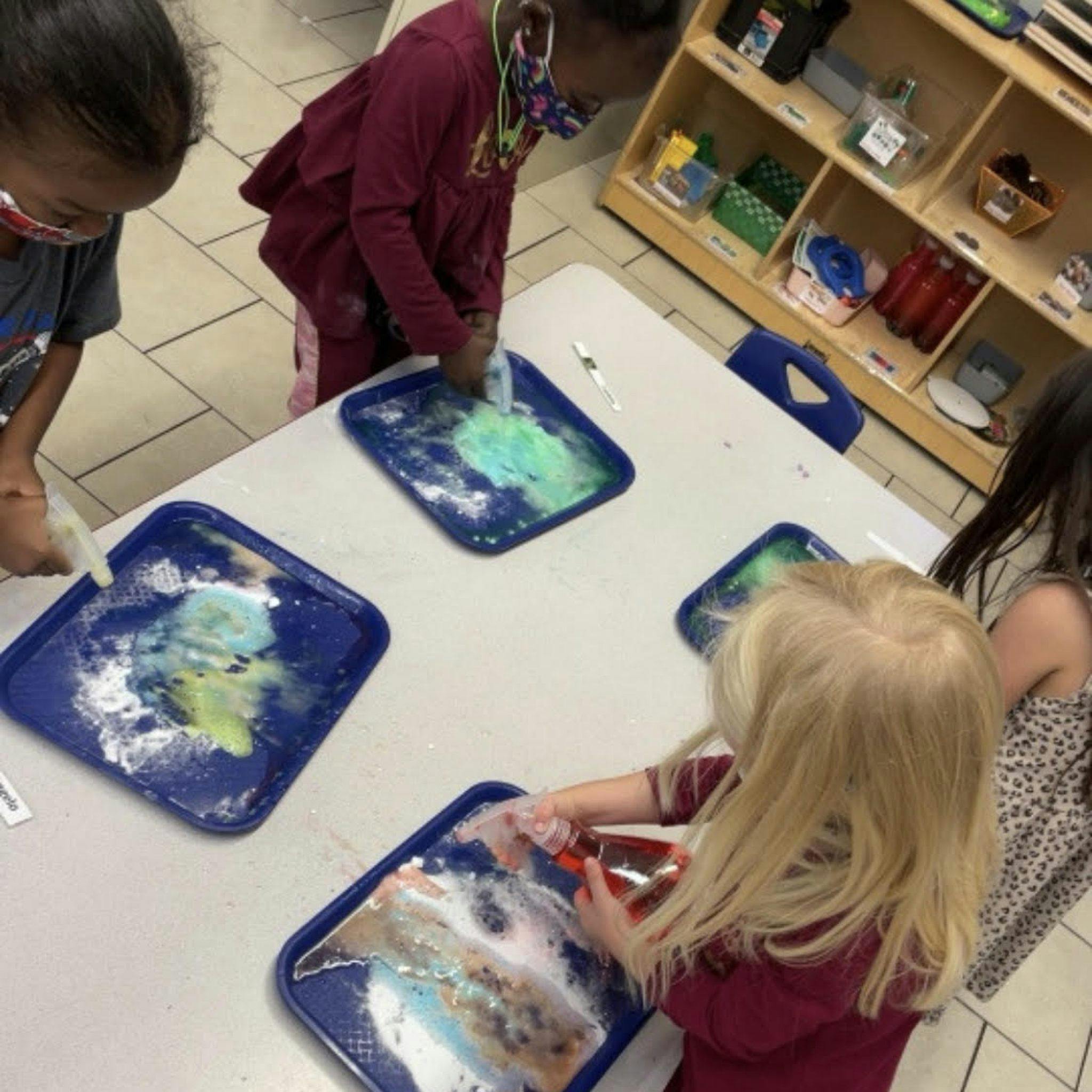 Children spraying liquids onto trays with colorful mixtures at a table