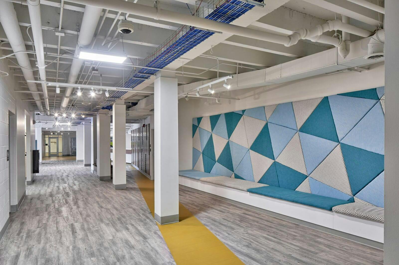Hallway with exposed ceiling pipes and a seating area with geometric wall panels