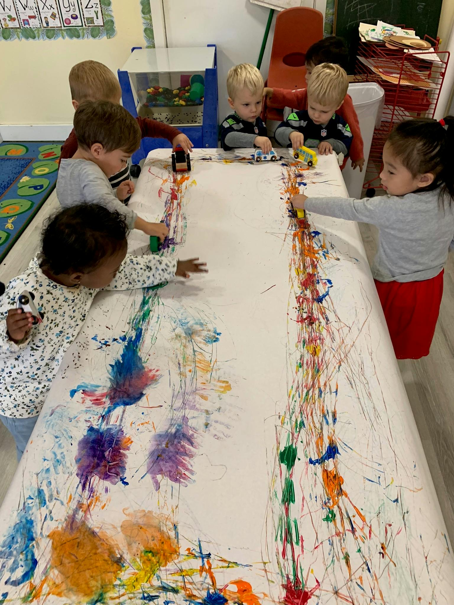 Children fingerpainting and playing with toy cars on a large table