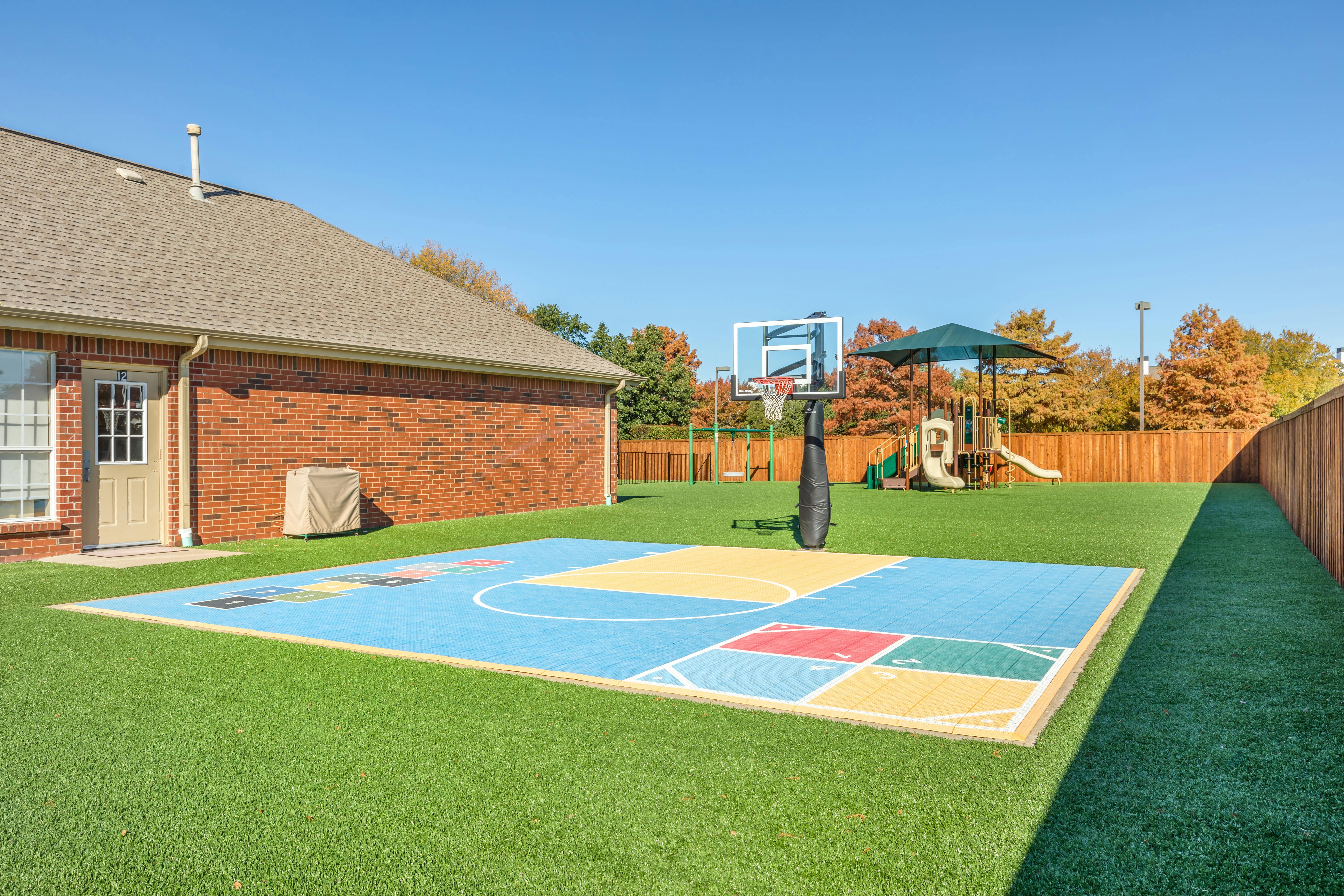 Basketball court on artificial grass next to a brick building and playground